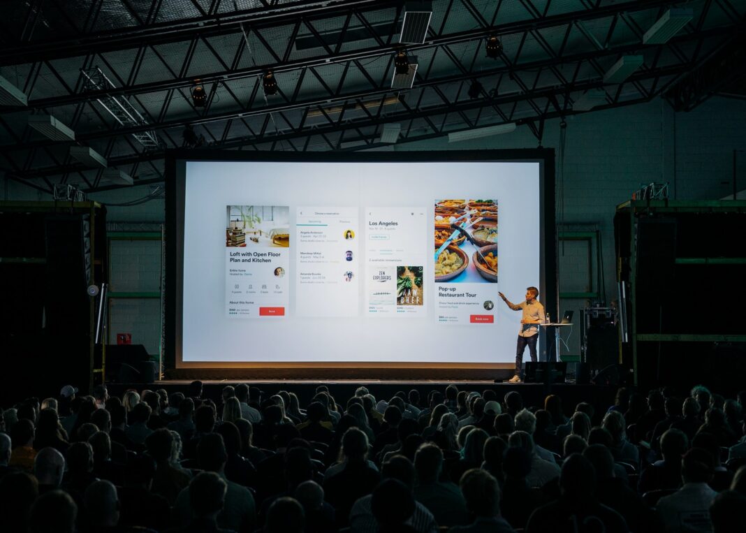 Photo by Teemu Paananen person discussing while standing in front of a large screen in front of people inside dim-lighted room