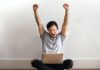 Spin and Win with These 10 Spectacular Sea-Themed Slots! Photo of Man in Gray T-shirt and Black Pants Sitting on Wooden Floor and Working on His Laptop Celebrating with His Hands Raised
