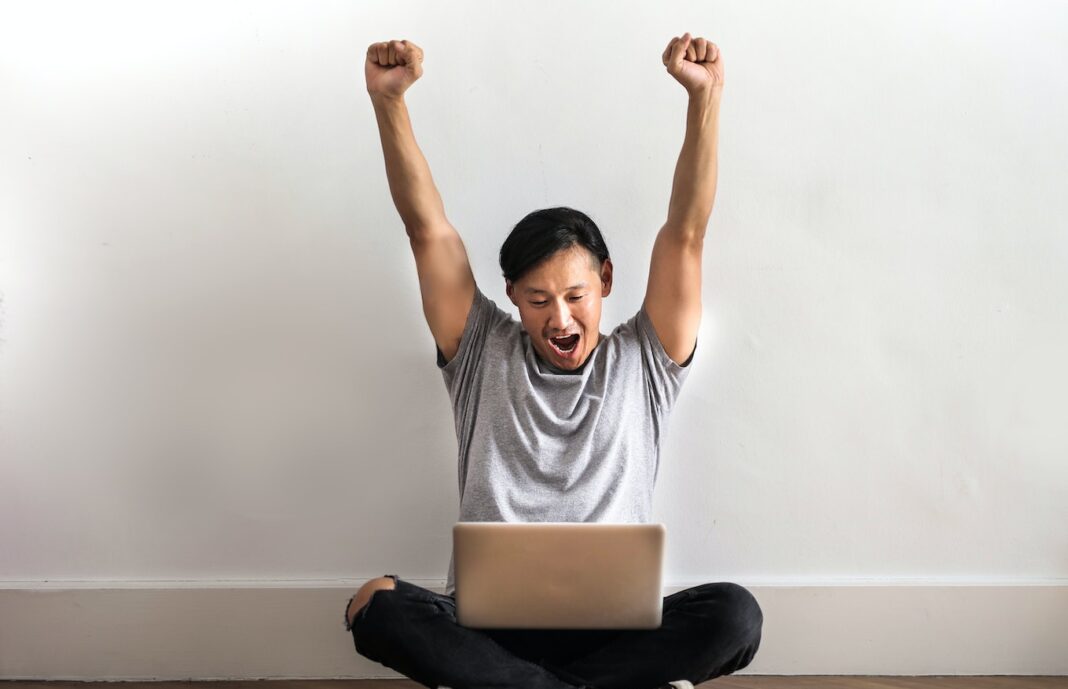 Photo of Man in Gray T-shirt and Black Pants Sitting on Wooden Floor and Working on His Laptop Celebrating with His Hands Raised