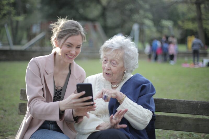 Photo by Andrea Piacquadio Delighted female relatives sitting together on wooden bench in park and browsing mobile phone while learning using