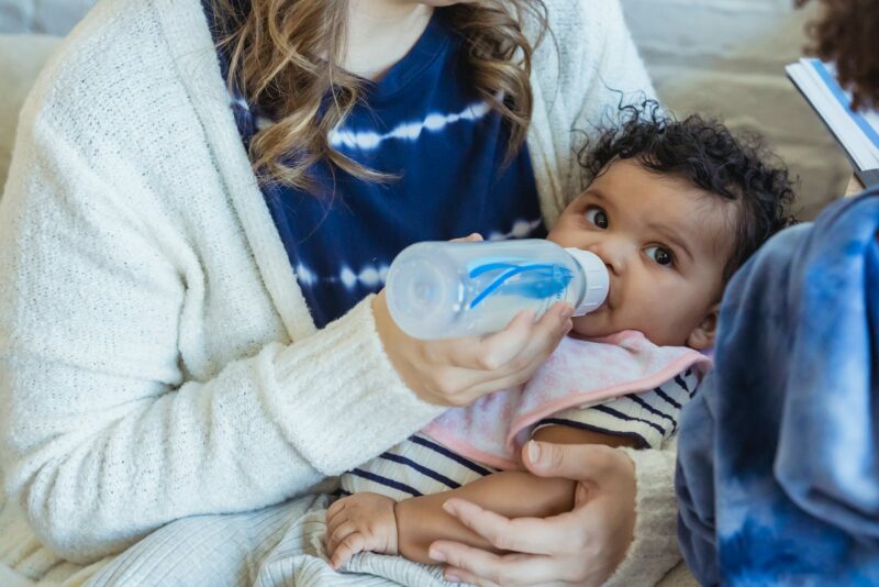 Photo by Keira Burton High angle of crop mother embracing little African American baby and feeding from bottle sitting on sofa near son