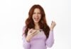 Satisfied redhead woman celebrating victory, say yeah yes and fist pump, winning on mobile phone, hold smartphone horizontally, standing against white background