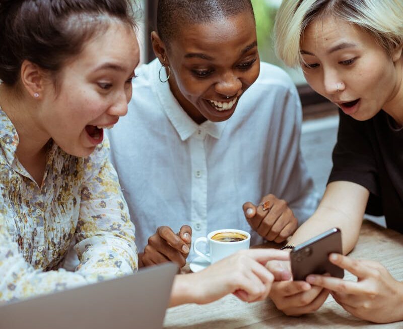 Photo by Ketut Subiyanto Amazed surprized multiethnic female friends in casual clothes with cup of coffee browsing smartphone while sitting at wooden table with laptop against window