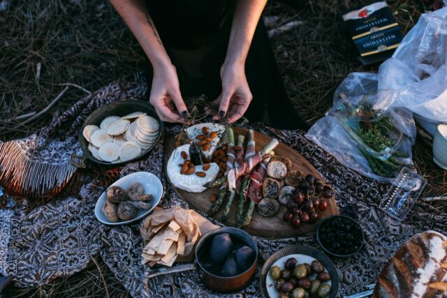 Photo by Rachel Claire From above crop anonymous female serving yummy food including asparagus with bacon potatoes and grapes on wooden board placed on plaid during picnic in nature