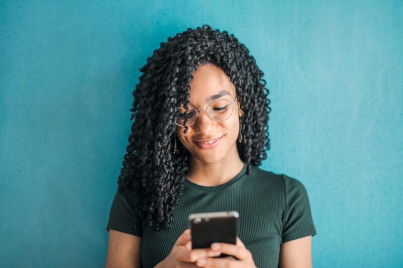 Photo by Andrea Piacquadio Portrait of a smiling woman using her phone, wearing eyeglasses, against a vibrant blue background.