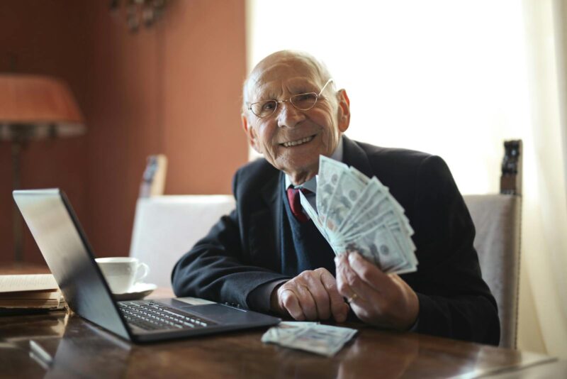 Photo by Andrea Piacquadio Happy senior businessman showing cash at his desk with a laptop, indoors.