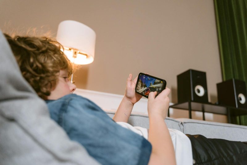 Photo by Tima Miroshnichenko Teen lying on sofa playing video games on smartphone indoors, surrounded by modern décor.