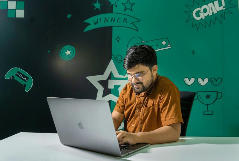 Photo by Ofspace LLC, Culture A man with a beard and glasses smiles while gaming on a laptop indoors, decorated with gaming graphics.