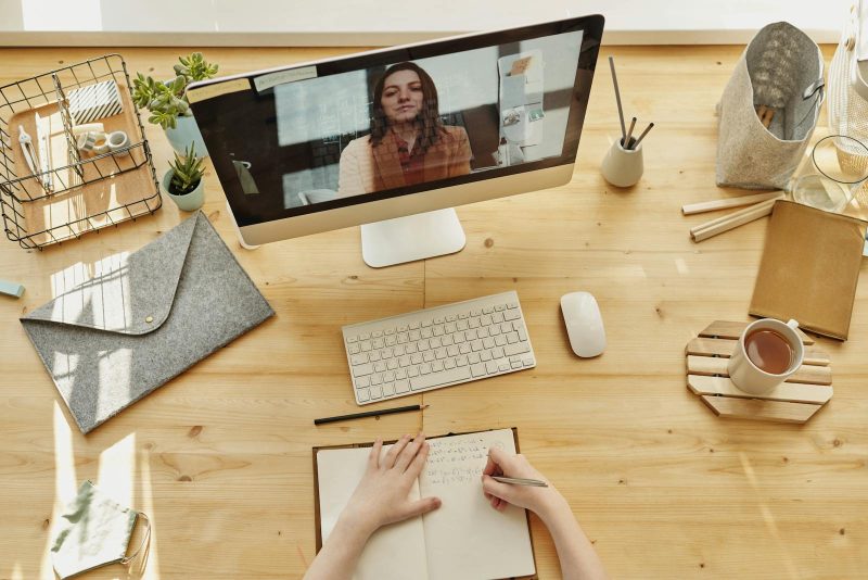 Photo by Julia M Cameron Person taking notes during a video call at a neatly organized home desk, showing remote work lifestyle.