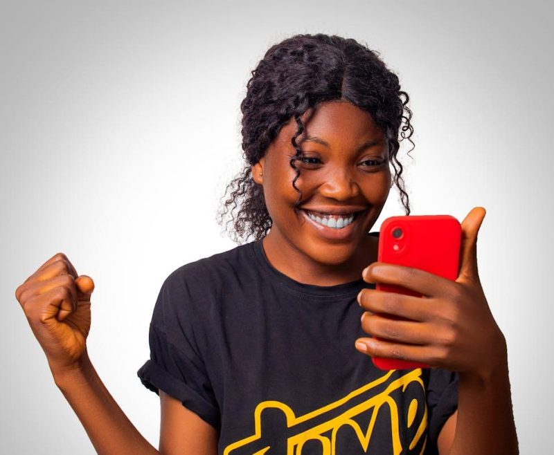 Photo by Ray Fotografy A cheerful woman celebrates success while looking at her smartphone, expressing happiness.