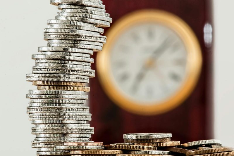 Photo by Pixabay A close-up image of stacked coins with a blurred clock, symbolizing time and money relationship.