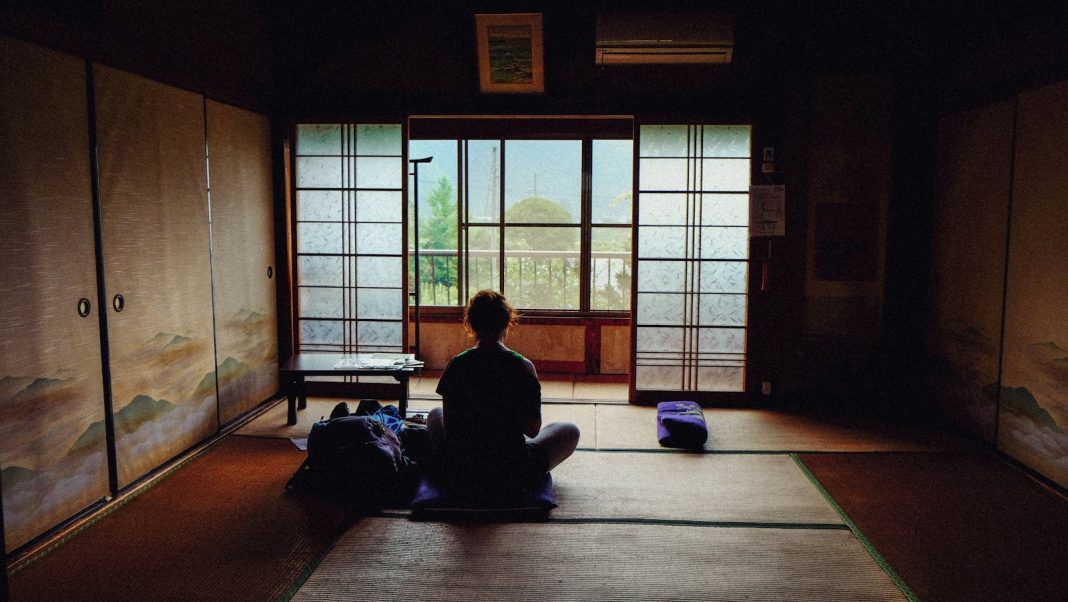 A person meditating in a tranquil traditional Japanese room, facing the view outside.