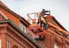 The ROI of a Solid Roof: Why Preventative Roofing Maintenance Pays Off for Commercial Properties Roofer in a cherry picker conducting maintenance on a red brick building with a tiled roof.