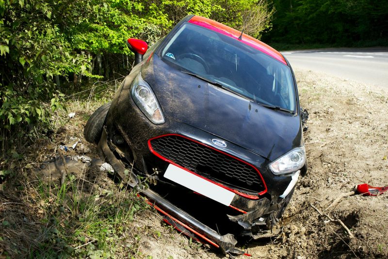 Photo by Mike Bird A black car, seemingly a Ford, crashed and damaged in a roadside ditch with surrounding greenery.