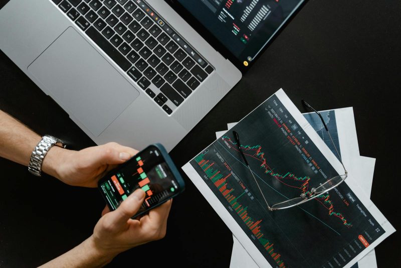 Photo by Tima Miroshnichenko Hands using smartphone beside laptop with stock charts, showcasing digital trading.