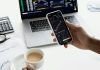 What Is Profit Storm? A person trades stocks on a smartphone with a laptop, coffee cup, calculator, and documents in view.