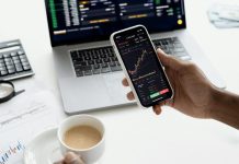 What Is Profit Storm? A person trades stocks on a smartphone with a laptop, coffee cup, calculator, and documents in view.