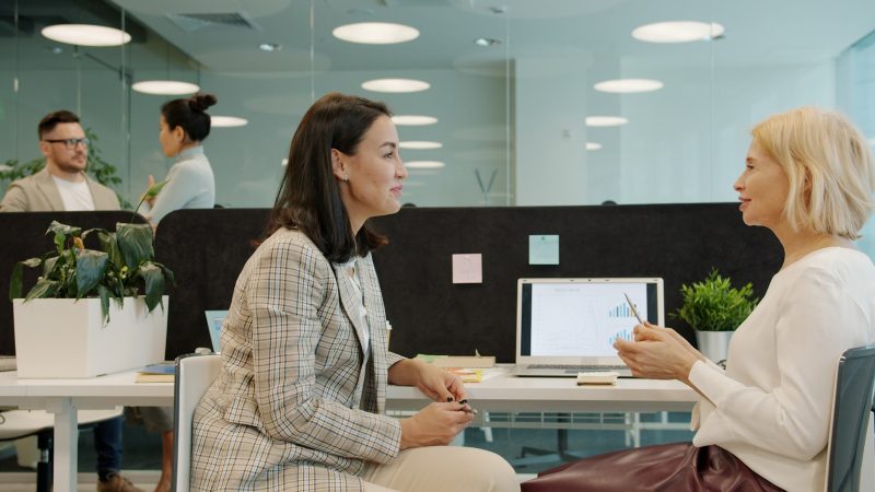 Photo by Vitaly Gariev Two women talking at a desk in an office.