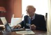 Senior man in formal attire using a laptop and holding money, symbolizing financial success.