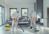 Two workers in protective gear vacuum and disinfect a modern living room.