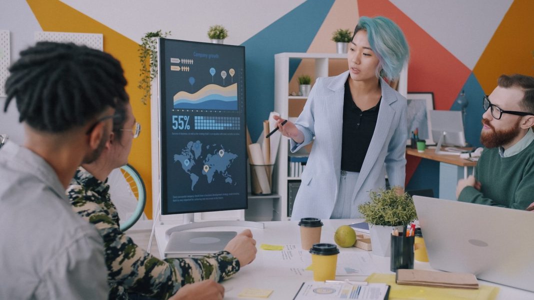 Photo by Vitaly Gariev A woman with blue hair presenting data on a monitor to a team